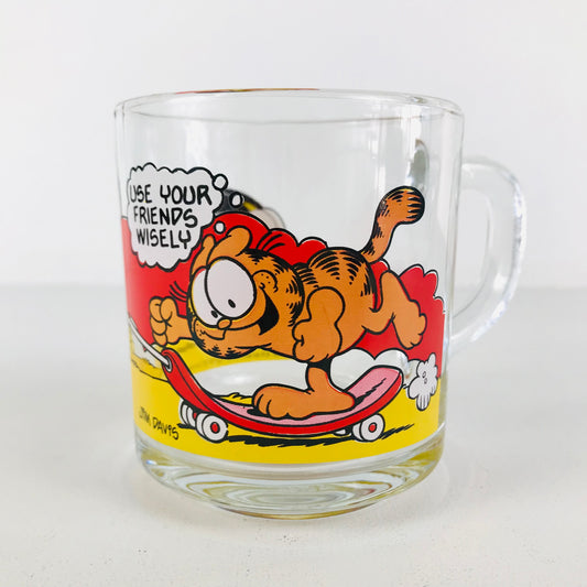 A glass mug with a comical image of Garfield riding on skateboard with a thought bubble that says "Use your friends wisely"