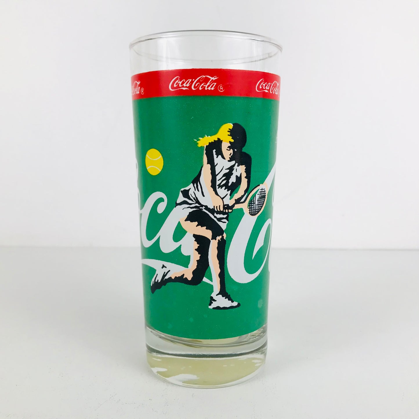 A vintage 1980s Coca Cola glass with a female tennis player swinging her racquet on the front.